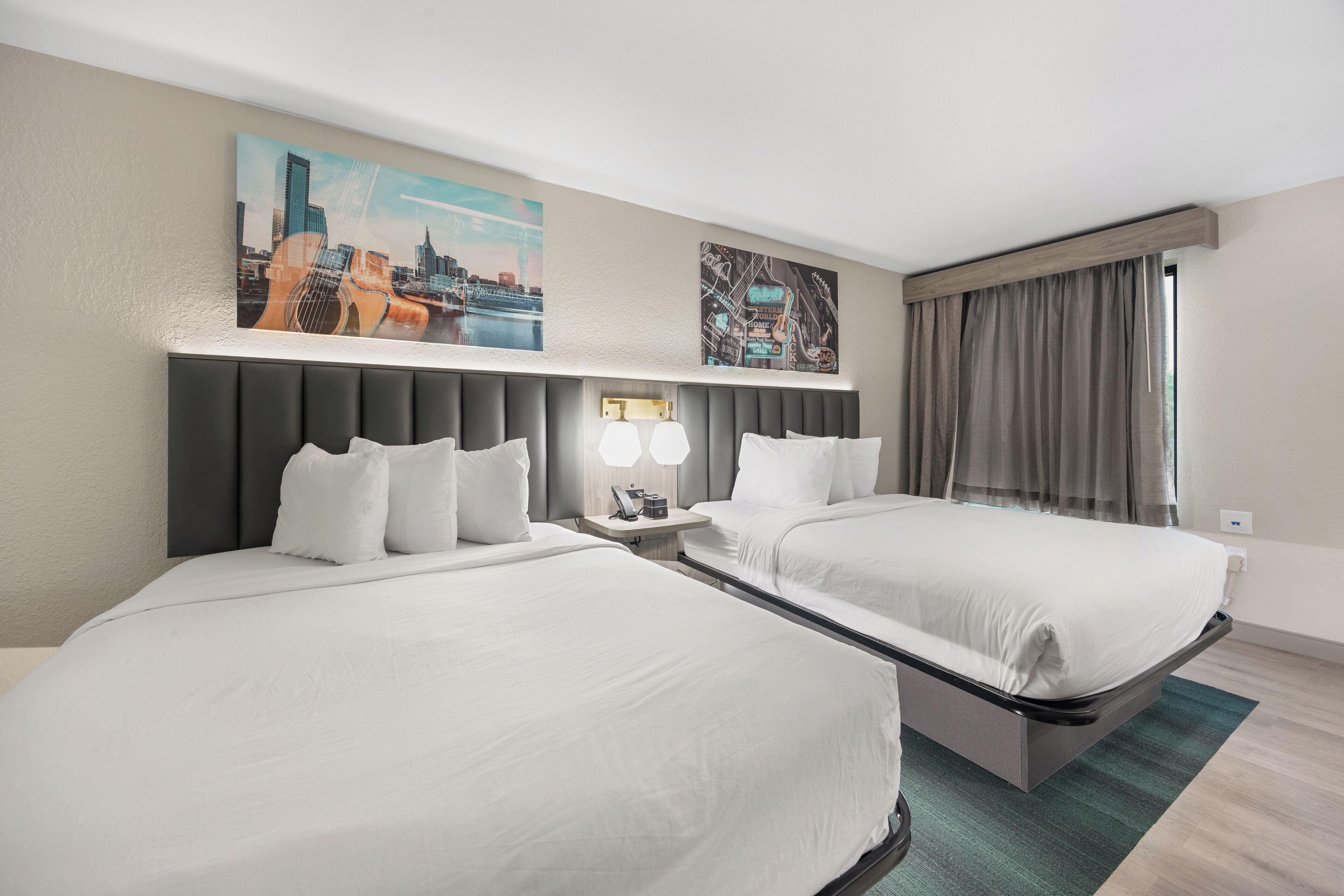 Habitación de hotel con dos camas, un cuadro de paisaje urbano, una mesita de noche con lámparas y una ventana con cortinas.