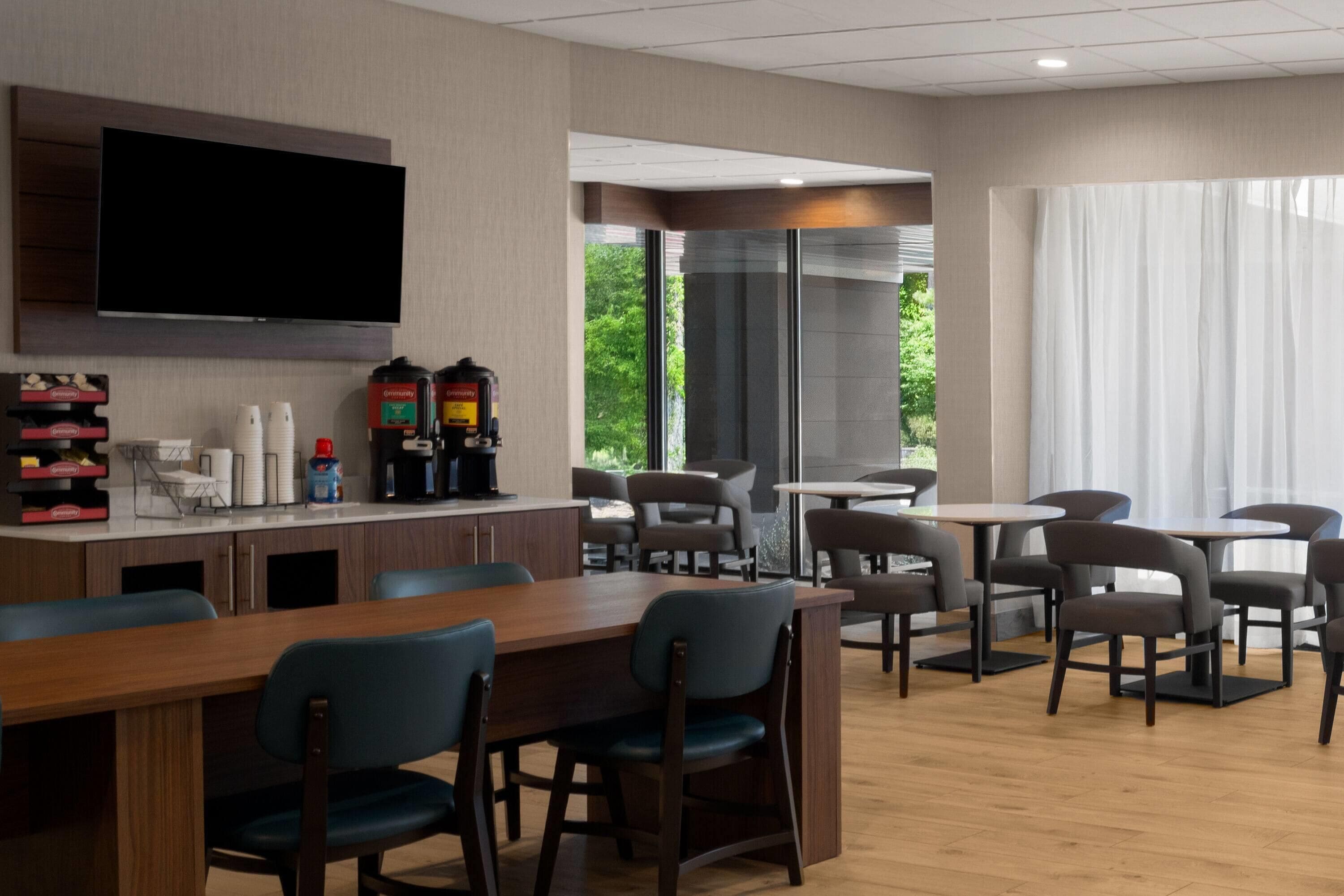 A modern hotel room with a flat-screen TV, a coffee machine, and a dining area with tables and chairs.