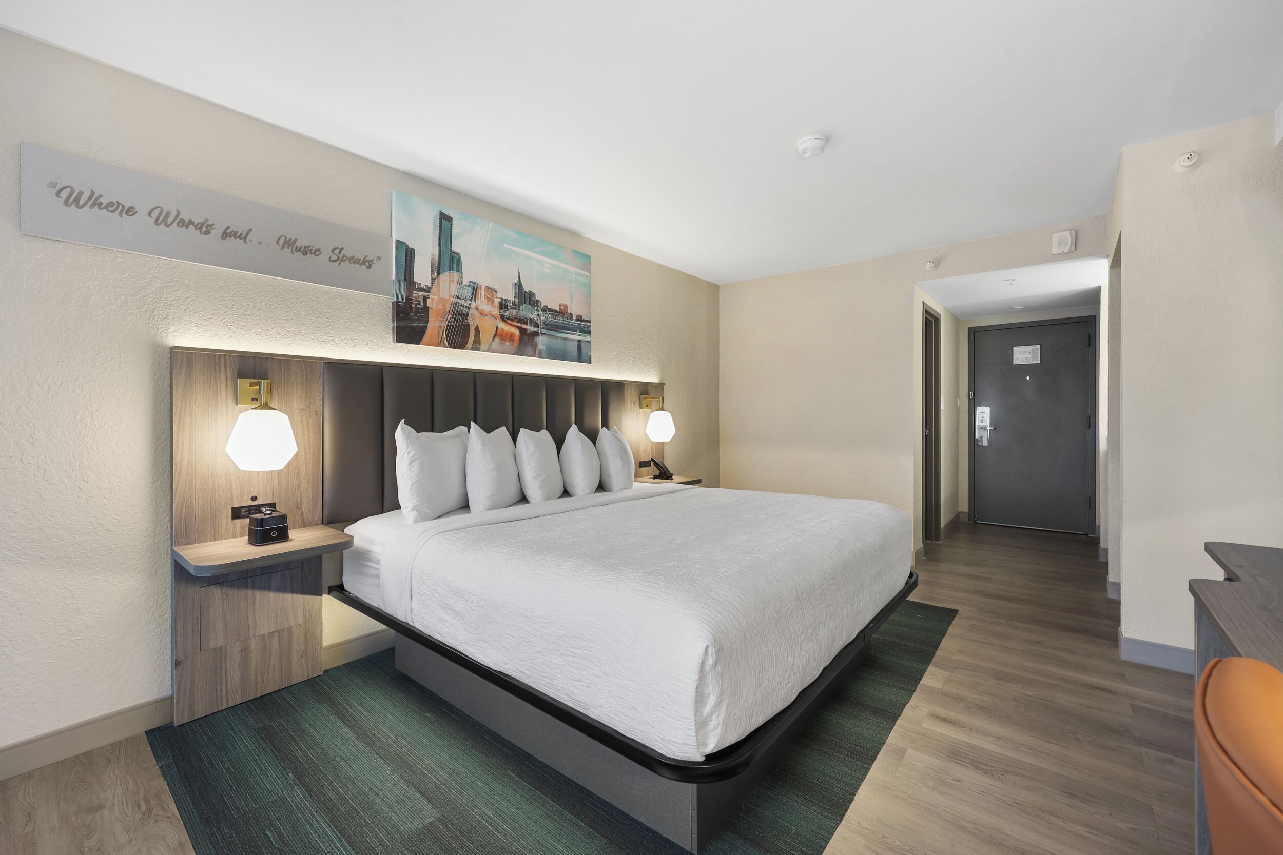 A hotel room with a bed, nightstands, a cityscape painting, a door, and a hallway.
