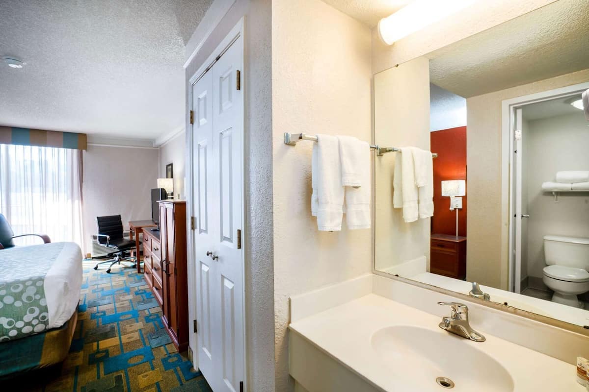 Quarto de hotel com cama, escrivaninha, cadeira e banheiro com pia e espelho.