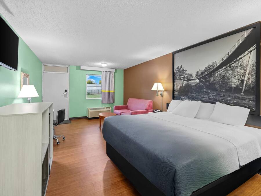 Quarto de hotel com cama, escrivaninha, cadeira, televisão e um quadro na parede.