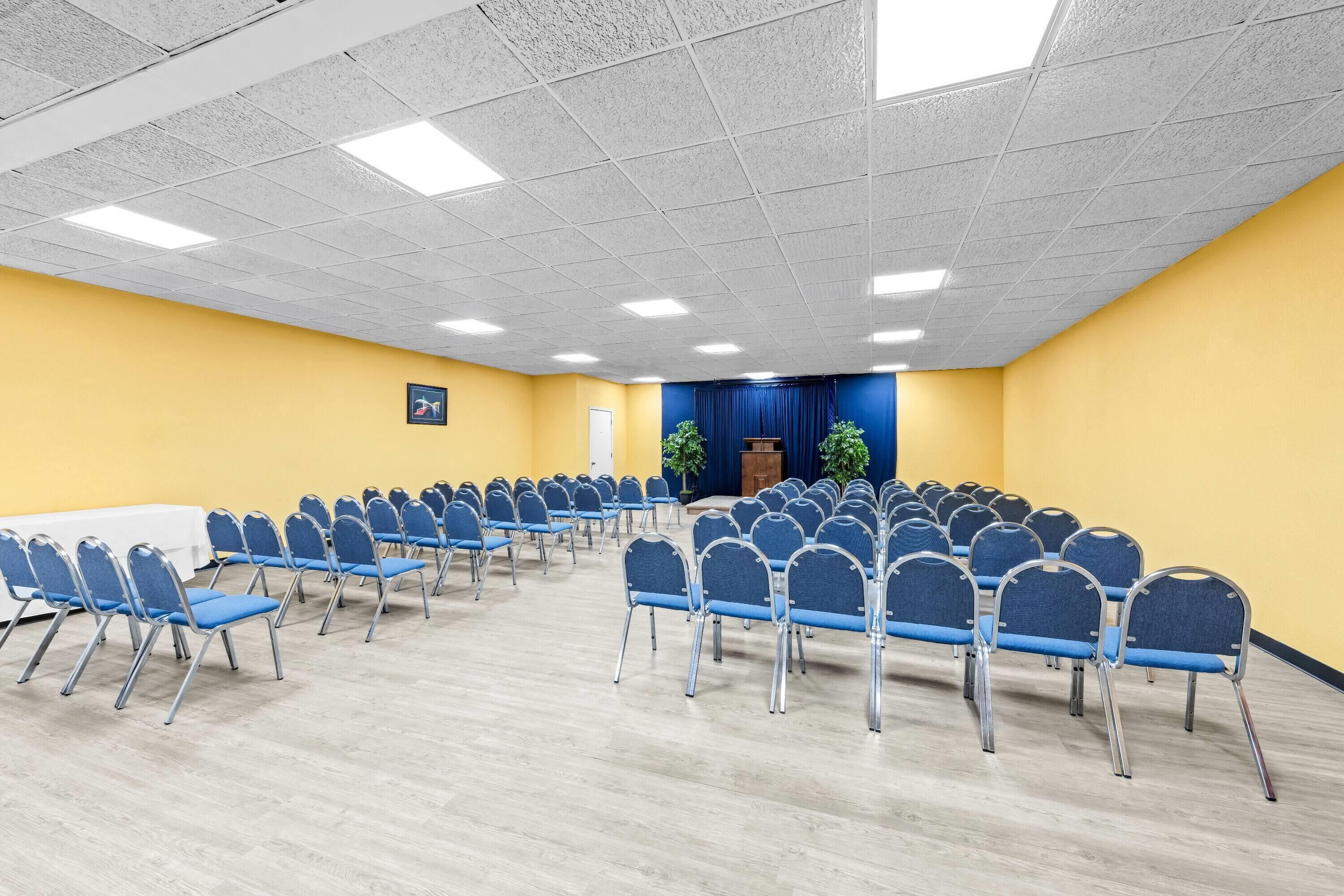 A conference room with rows of blue chairs, a podium, and a stage with a blue curtain.