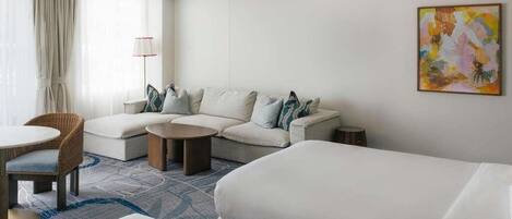 Une chambre d’hôtel avec un canapé, deux lits simples, une table basse ronde, une chaise et une lampe.
