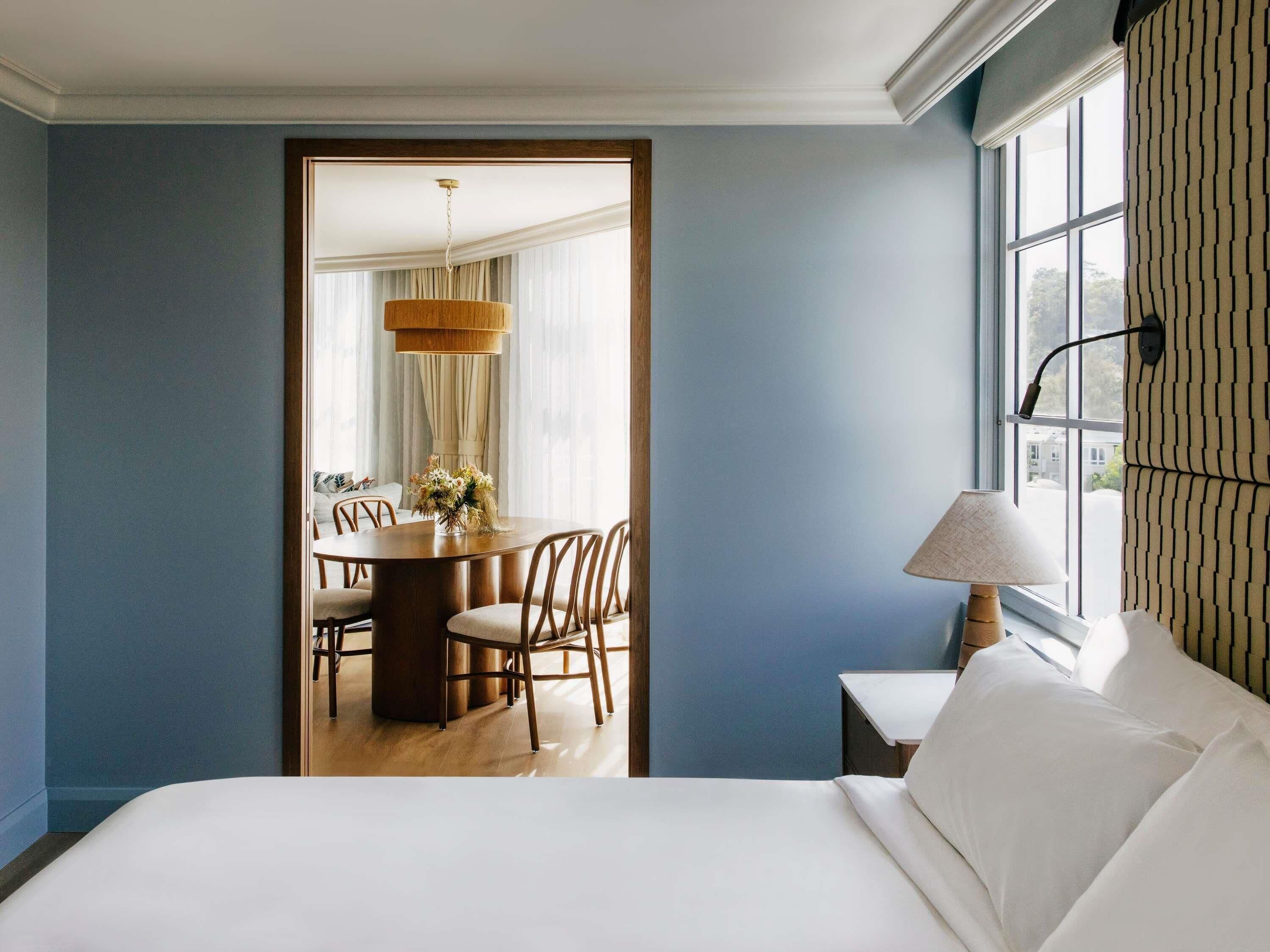A hotel room with a bed, a dining table with chairs, a lamp, and a window with curtains.