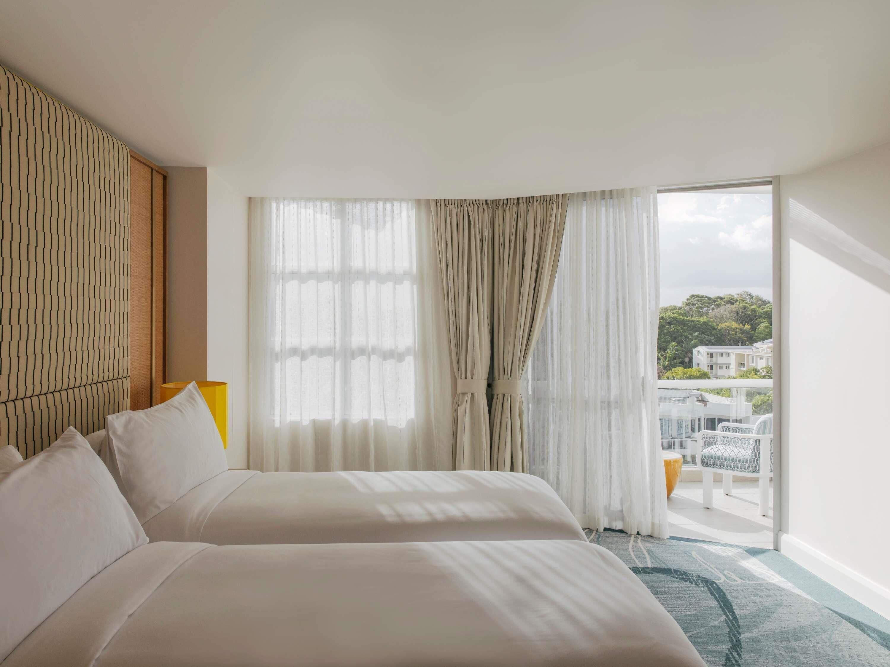 Habitación de hotel con dos camas, un ventanal con cortinas translúcidas, vistas al exterior y un balcón con mobiliario.
