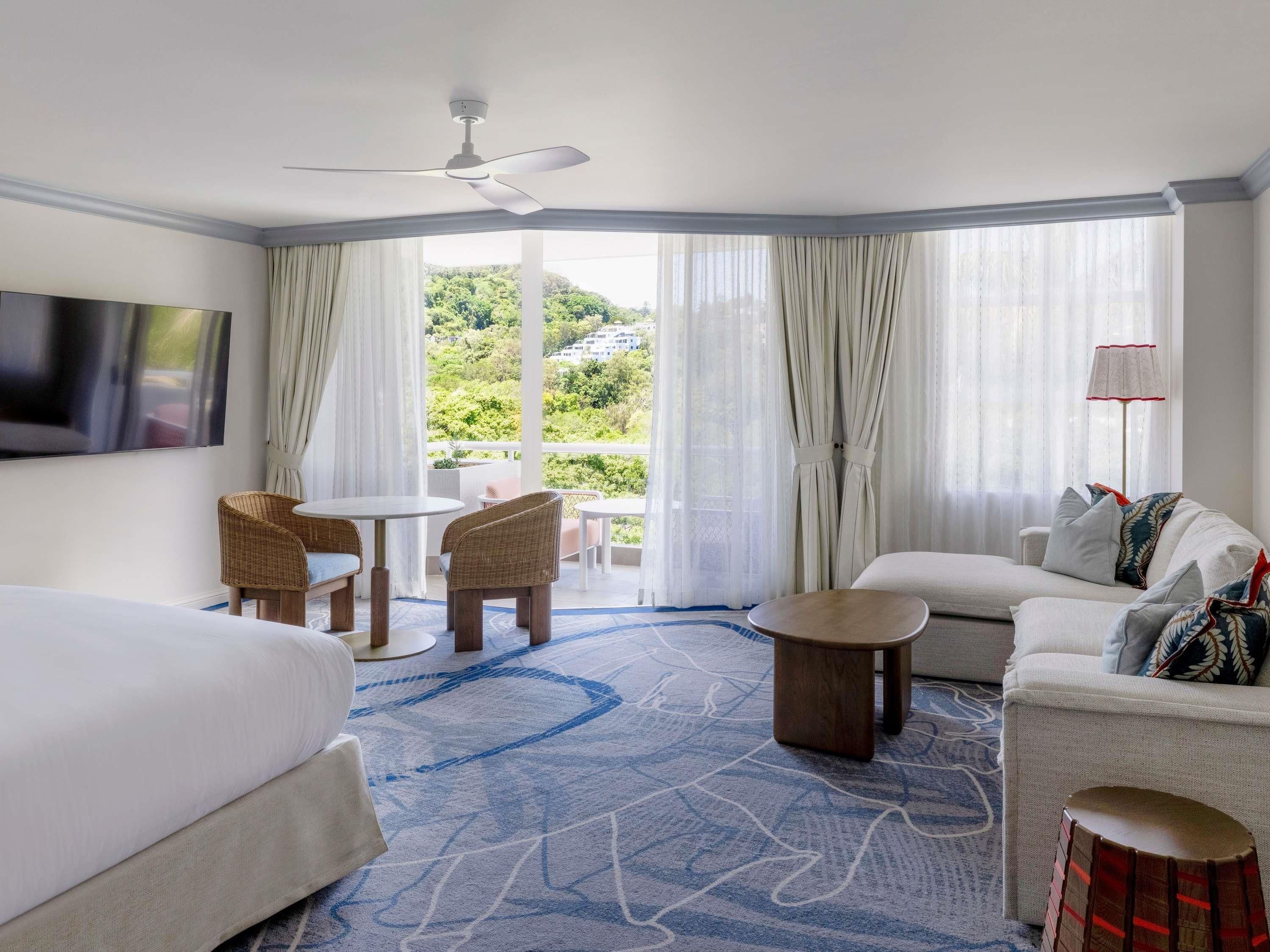 A hotel room with a bed, a sofa, a dining table with chairs, a ceiling fan, and a view of greenery outside.
