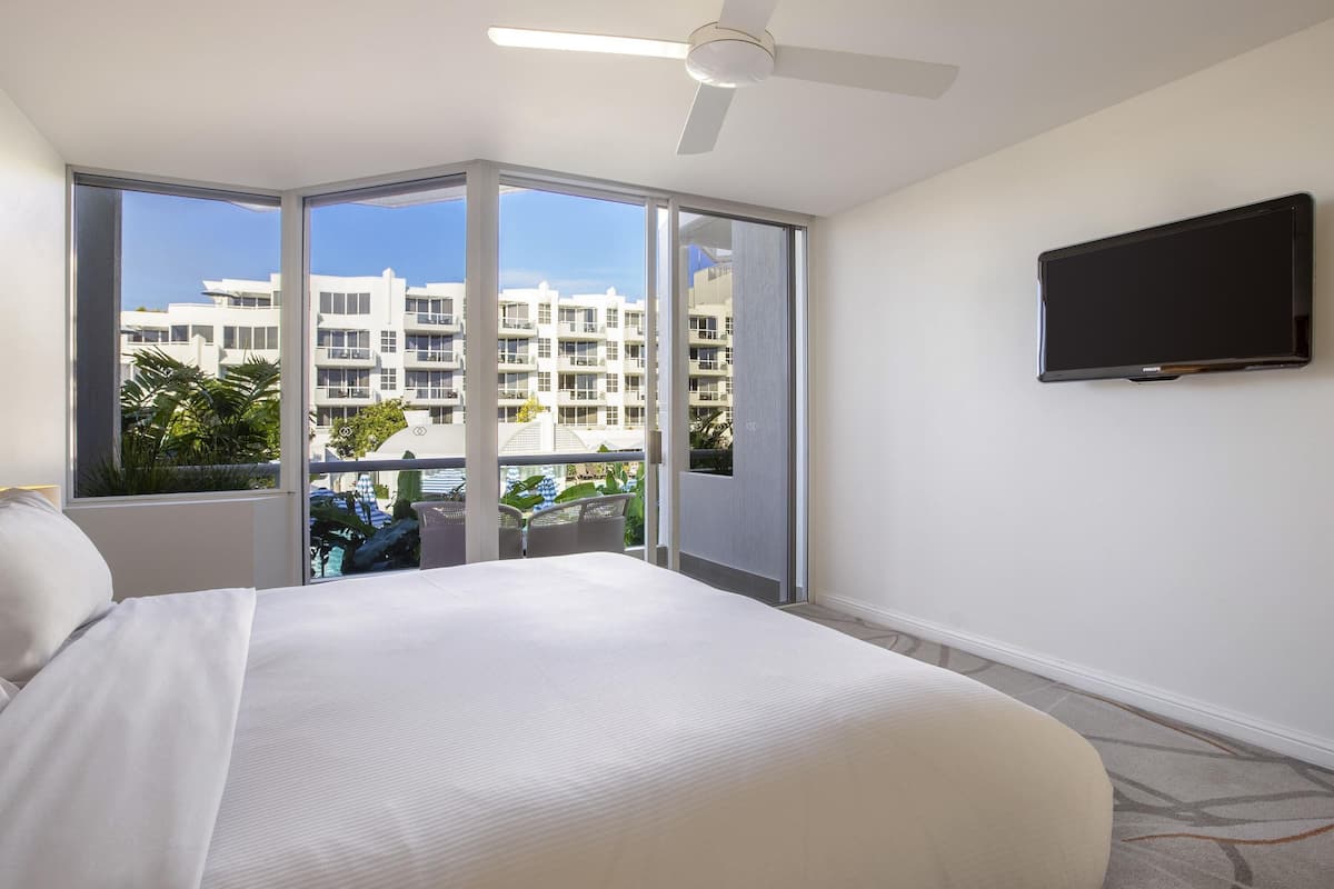 A hotel room with a bed, a ceiling fan, a TV, and large windows offering a view of a building and palm trees.