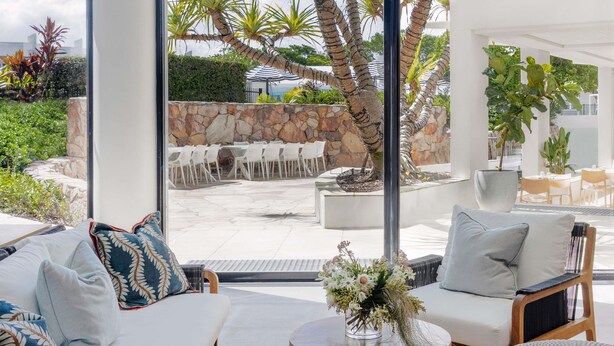 A modern outdoor seating area with white chairs and tables, a stone wall, and a large tree.