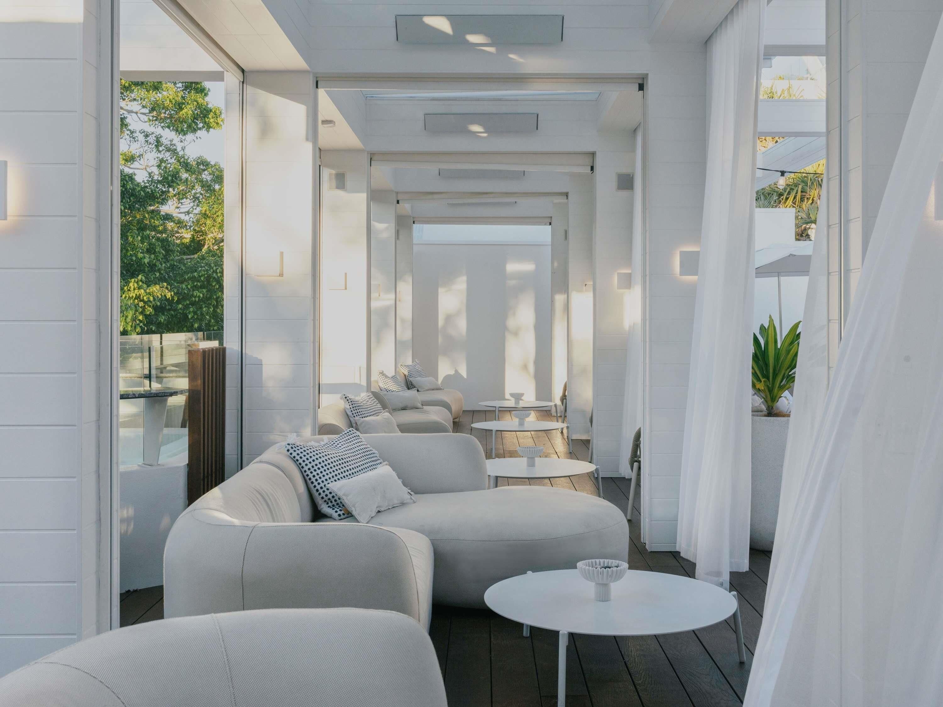 A modern outdoor seating area with a sofa and tables, enclosed by glass walls and white curtains.