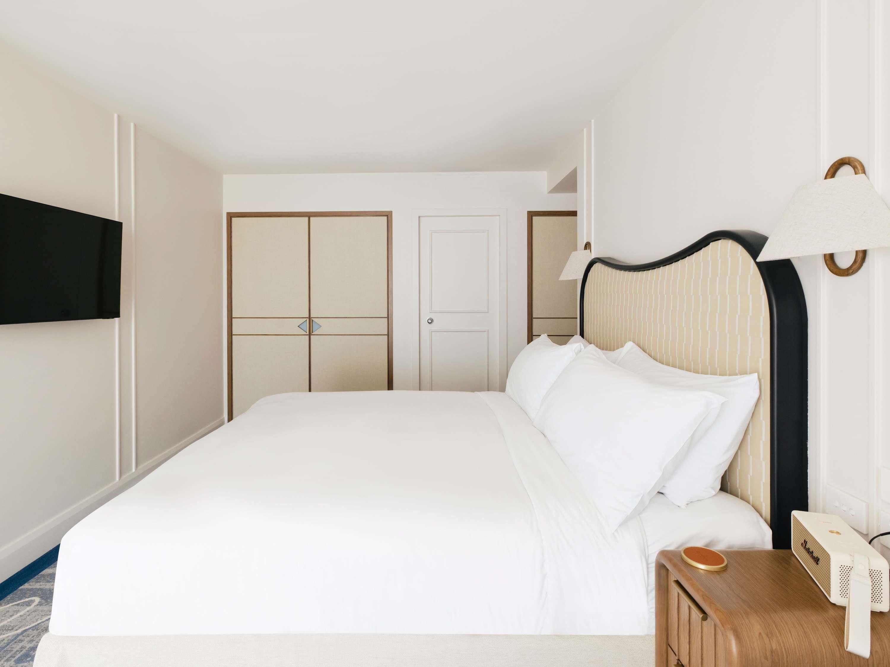 A hotel room with a large bed, a bedside table with a lamp, a flat-screen TV mounted on the wall, and a wardrobe with a mirror.