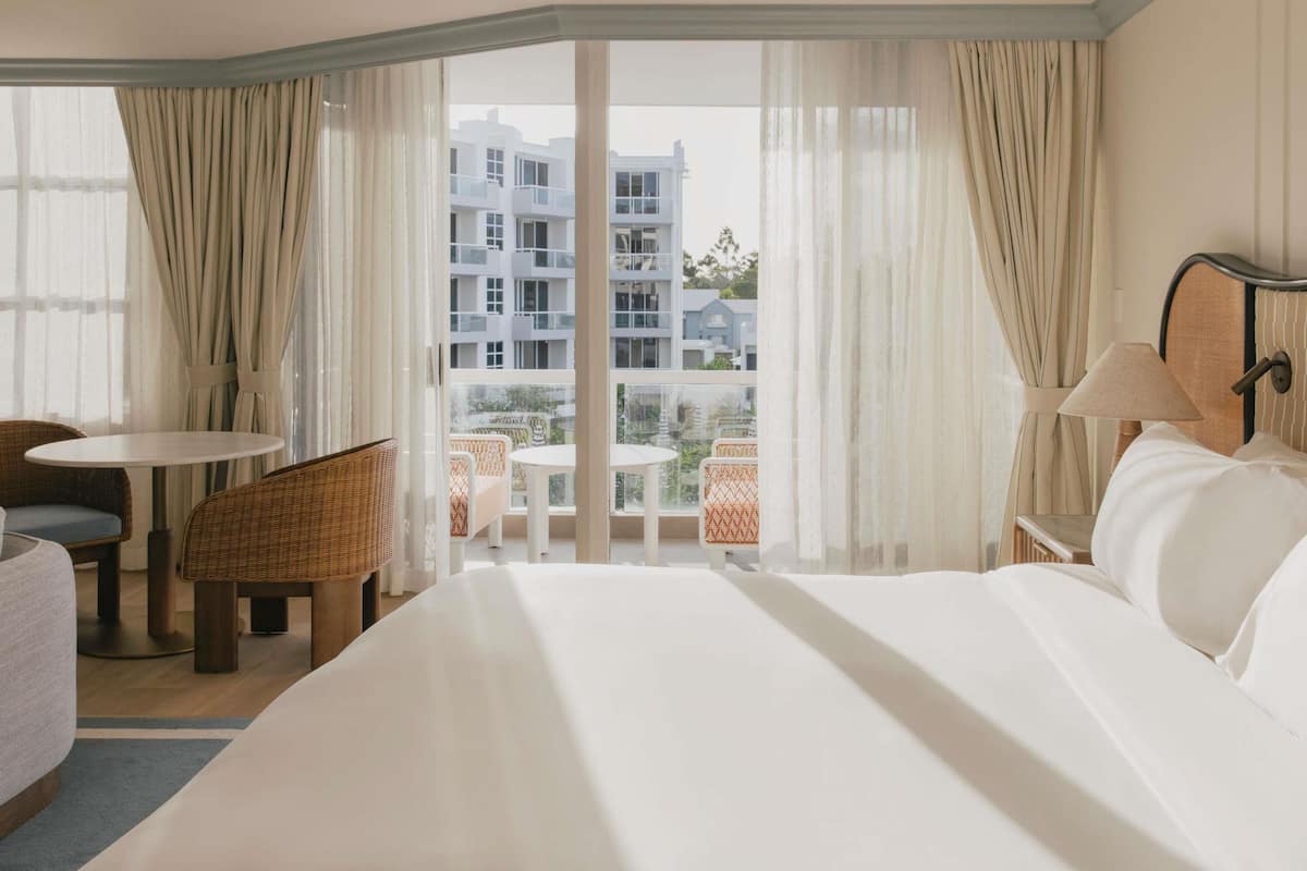 A hotel room with a large bed, a dining table, wicker chairs, and a view of a building complex.