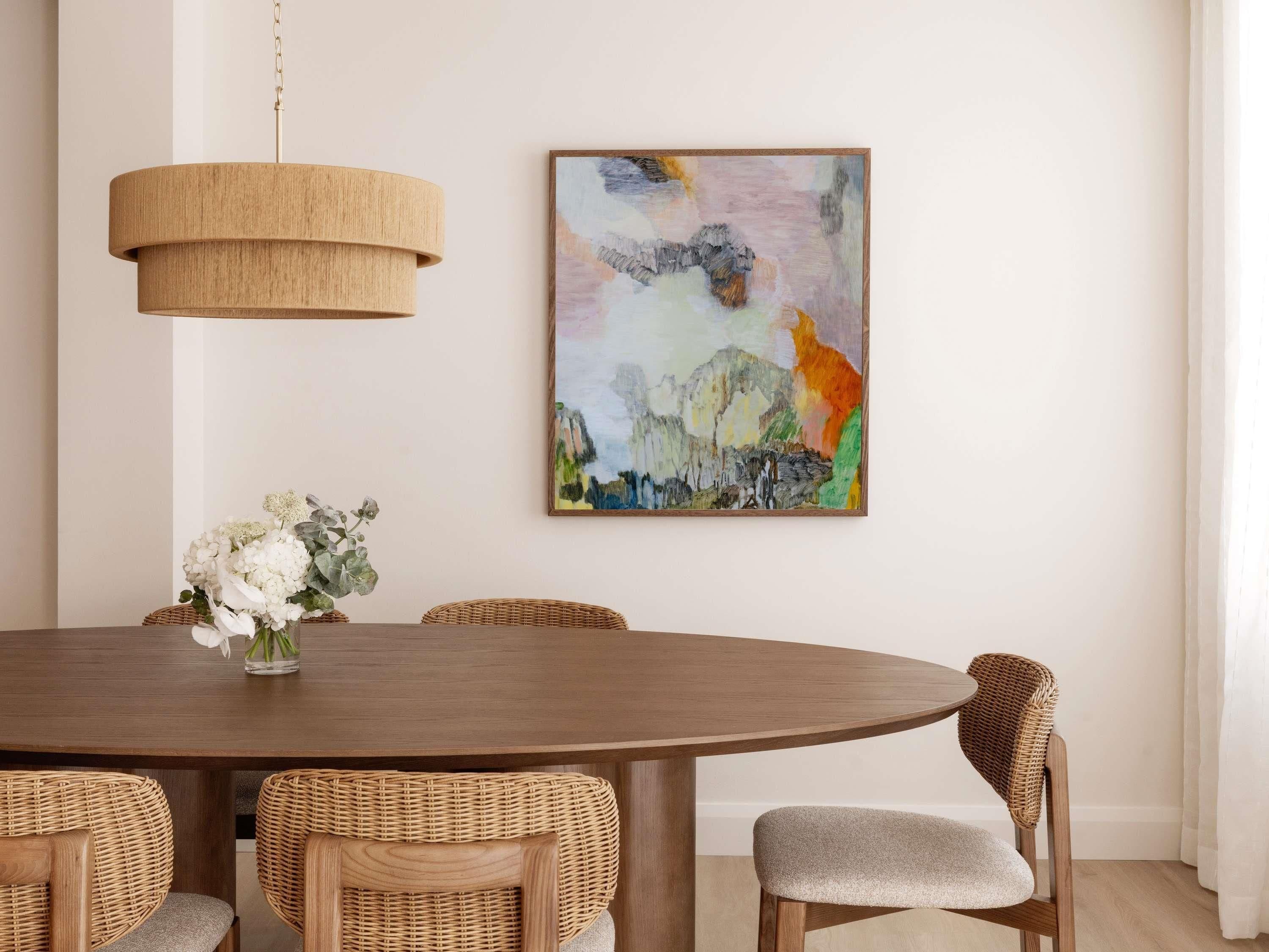 A dining area with a round table, wicker chairs, a hanging light fixture, and a colorful abstract painting on the wall.