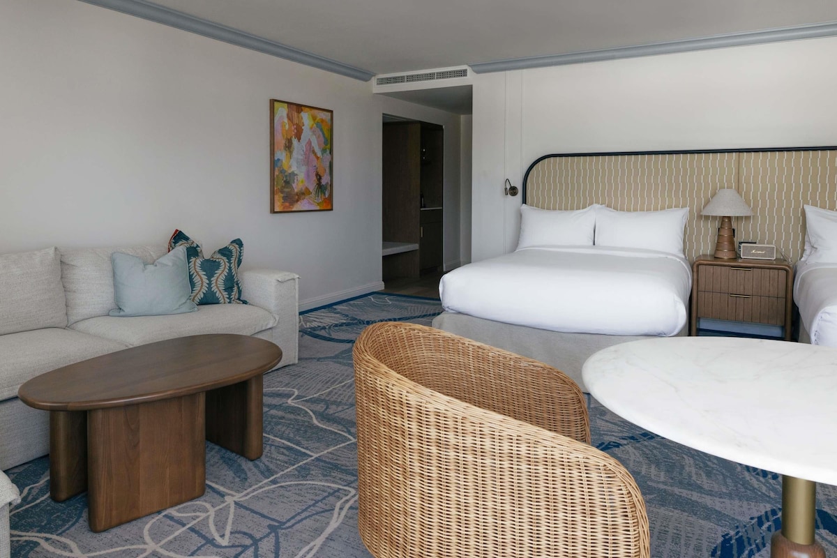 A hotel room with a bed, a sofa, a coffee table, a wicker chair, and a bedside table with a lamp.