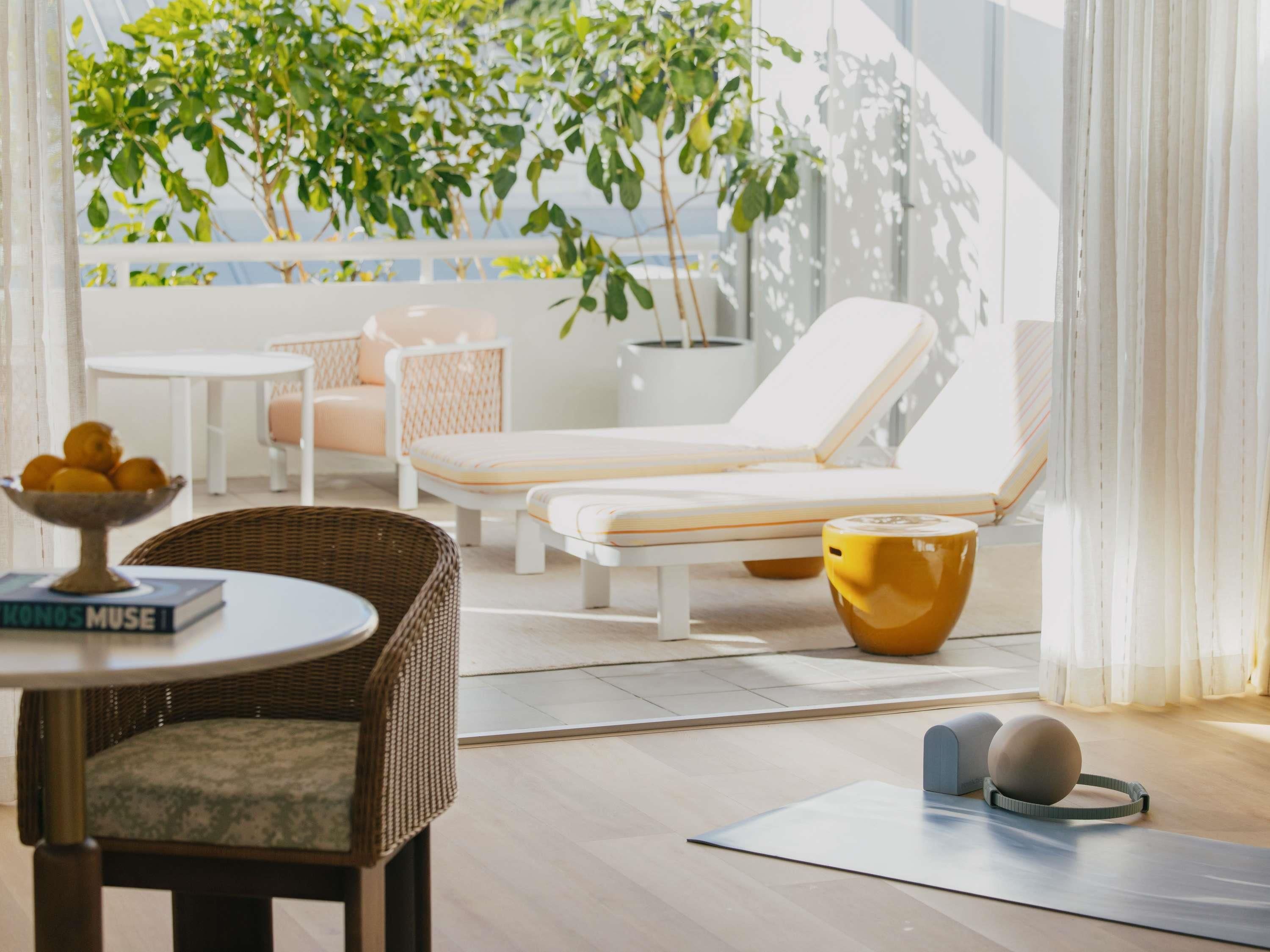 A modern lounge area with a white sofa, a wicker chair, a small table with a bowl of fruit, and a yoga mat.