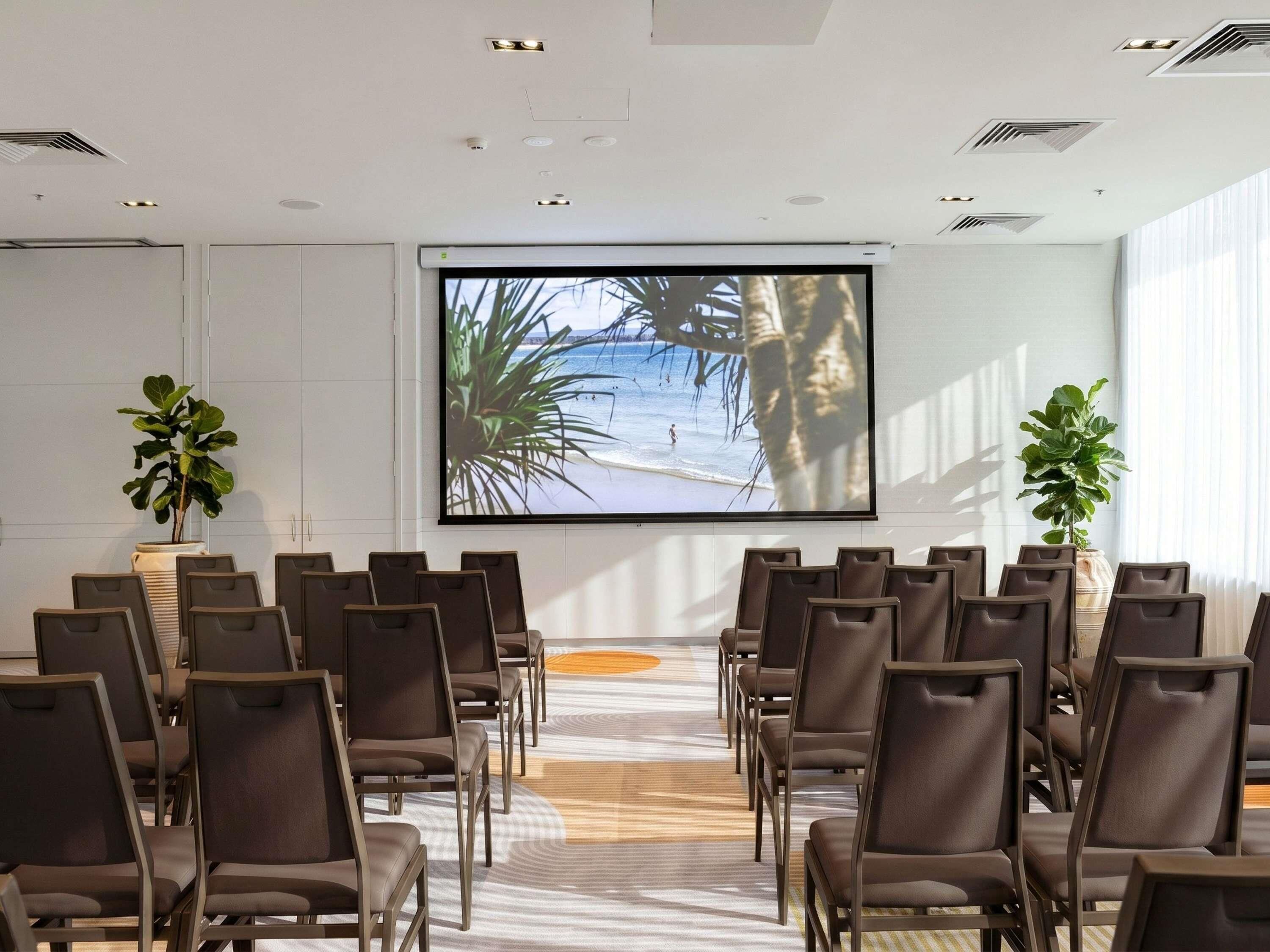 Un sal ón de conferencias con una pantalla grande mostrando una escena de playa, asientos dispuestos y plantas en macetas.