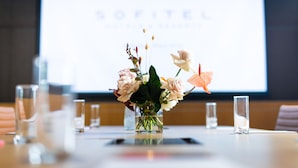 Una mesa de conferencias con un centro de mesa floral, vasos y una pantalla de proyección al fondo.