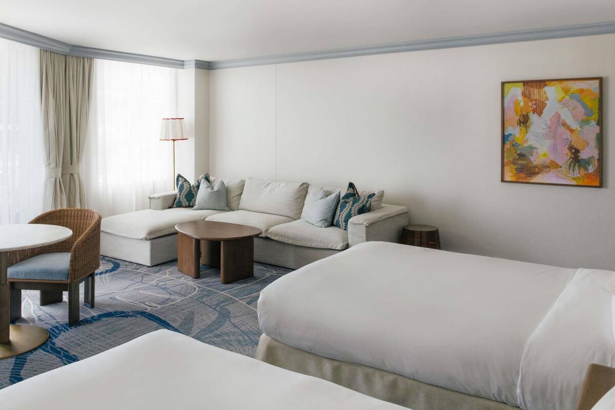 A hotel room with a sofa, two single beds, a round coffee table, a chair, and a lamp.