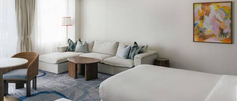 A hotel room with a sofa, two single beds, a round coffee table, a chair, and a lamp.