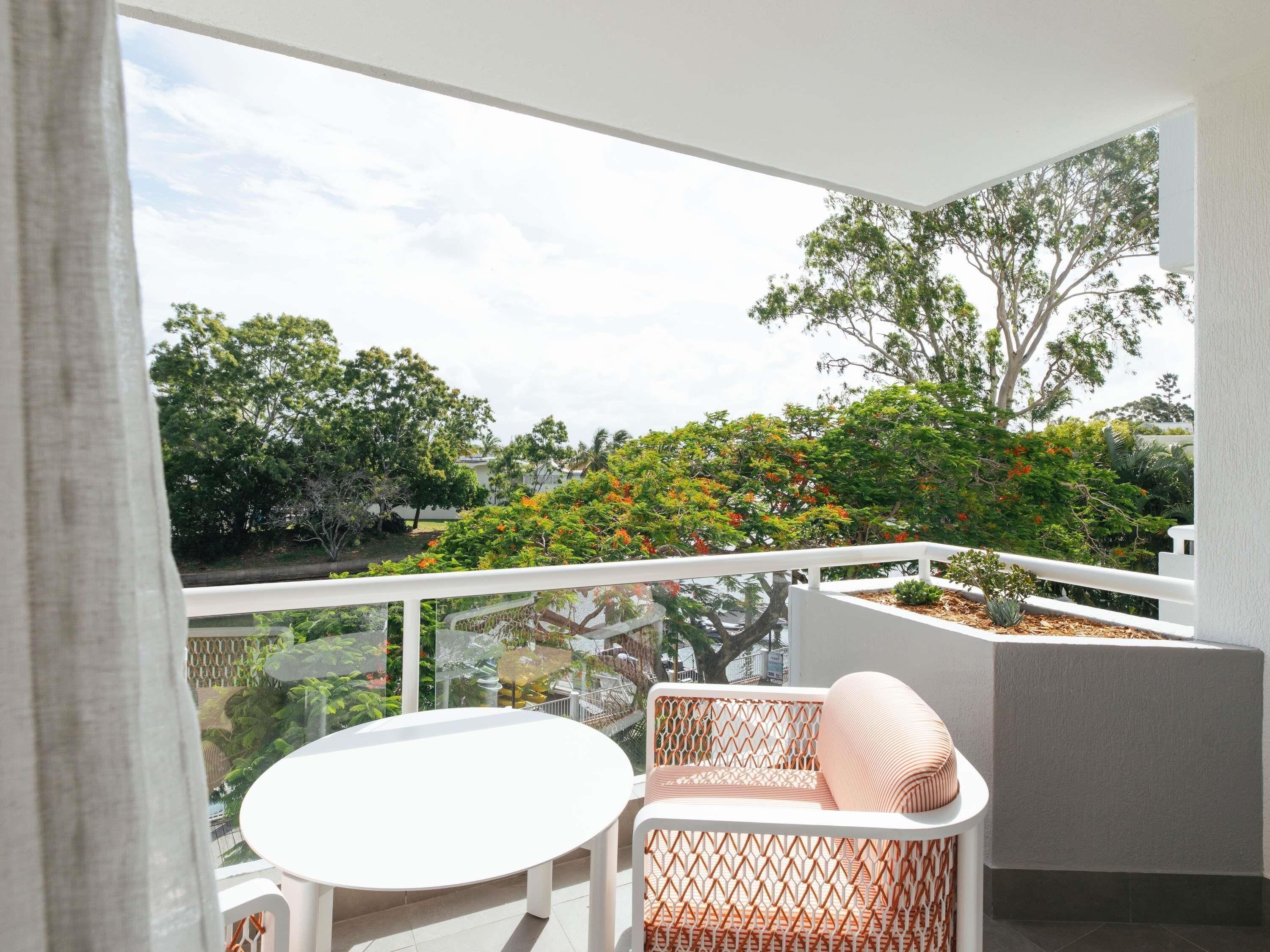 A balcony with a white table and wicker chair, overlooking a garden with trees and shrubs.