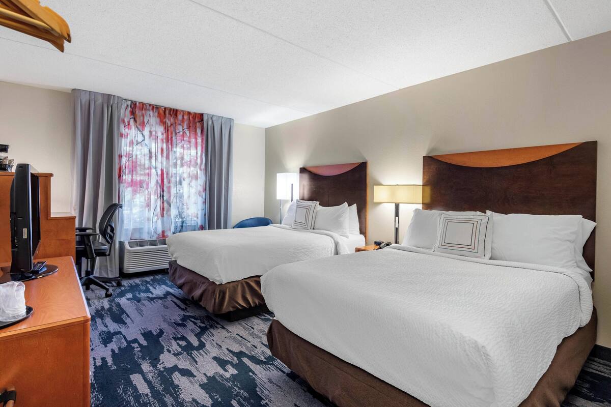 Una habitación de hotel con dos camas, un escritorio con computadora, una silla y una ventana con cortinas.