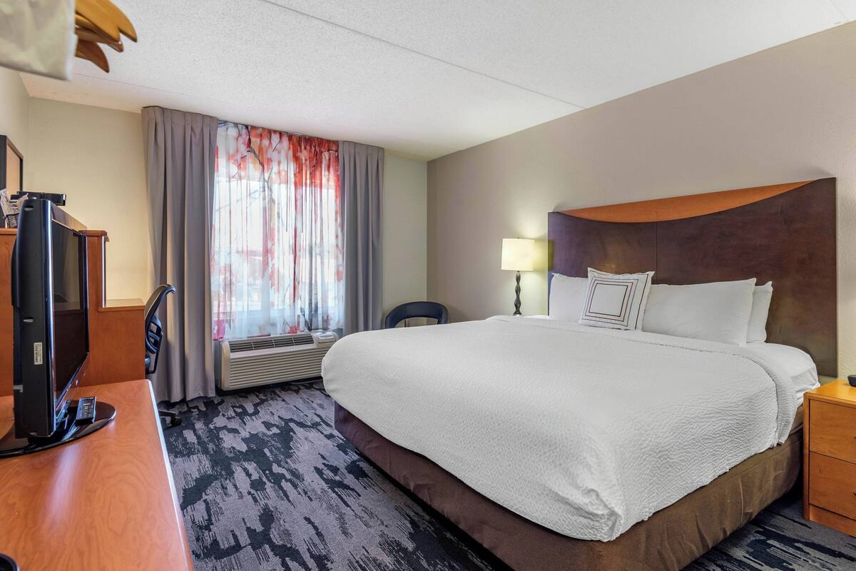 Habitación de hotel con una cama grande, un escritorio con computadora, una silla, una lámpara y un ventanal con cortinas.