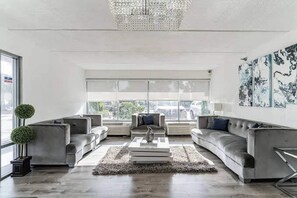 A modern living room with a chandelier, grey sofas, a coffee table, and a large window with a view. - Home 1 Suites Extended Stay - Kissimmee (Kissimmee)
