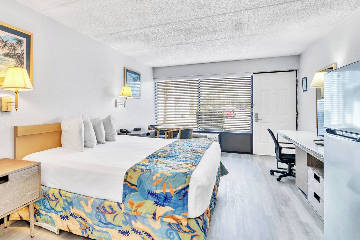 A hotel room with a bed, bedside table, desk, chair, and a window with blinds.