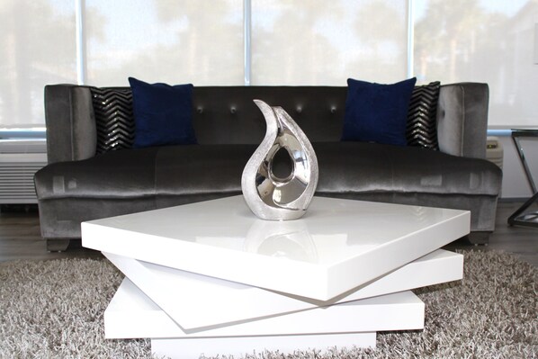 A modern living room with a sleek black sofa, two blue pillows, and a metallic vase on a white coffee table. - Home 1 Suites Extended Stay - Kissimmee (Kissimmee)