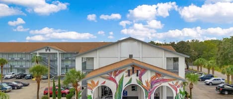 A hotel with a parking lot, a covered entrance with colorful plant murals, and a multi-story building in the background.