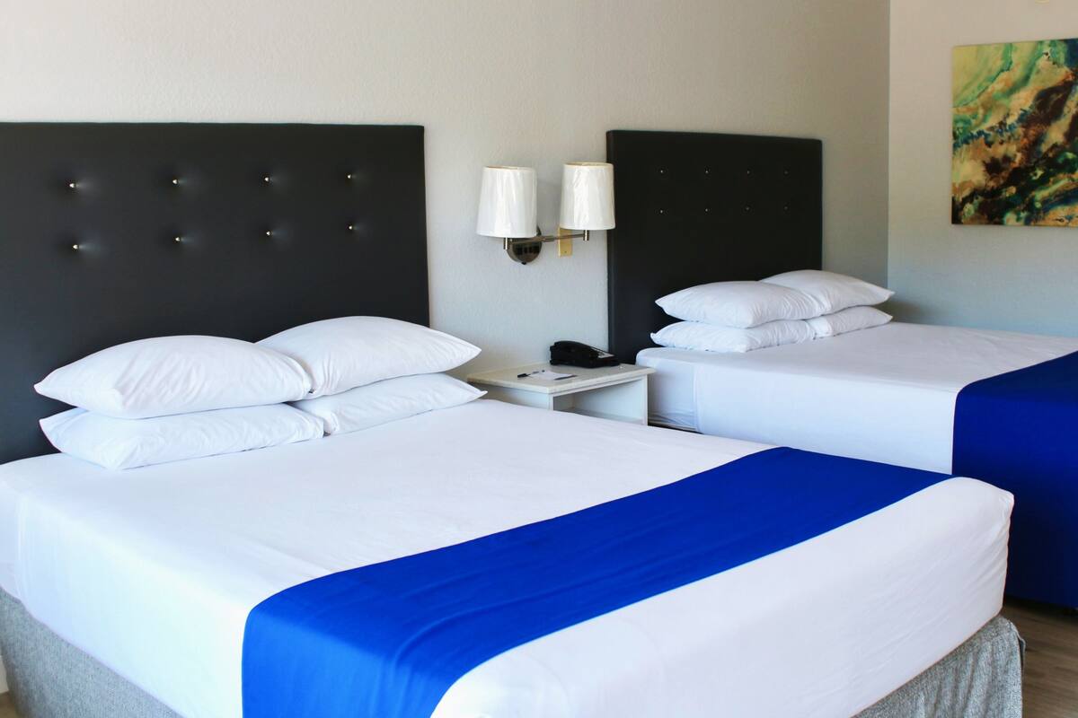 A hotel room with two beds, each with white bedding and a blue accent. There are two bedside tables with lamps, and a painting on the wall.
