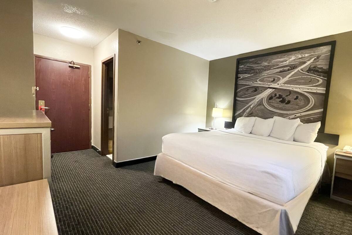 A hotel room with a large bed, two bedside tables with lamps, a desk with a chair, a door, and a wall with a road map mural.