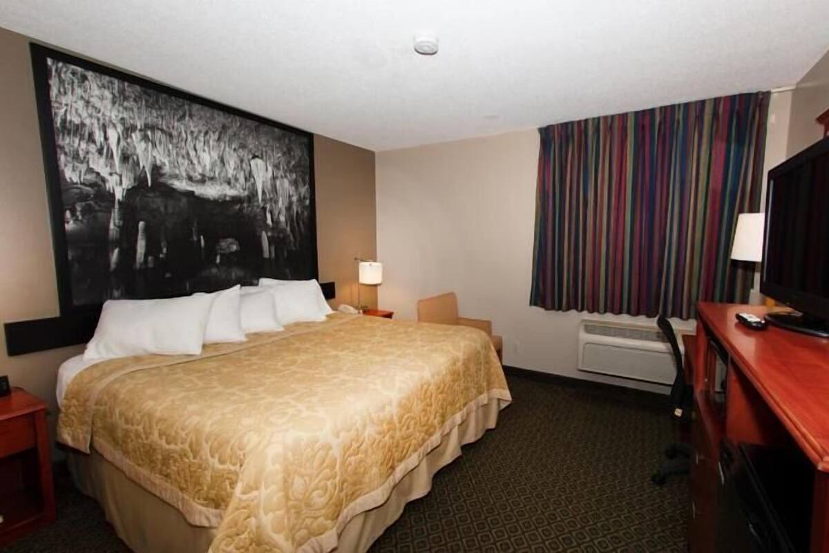 A hotel room with a bed, a desk with a computer, a television, a large painting on the wall, and a window with curtains.