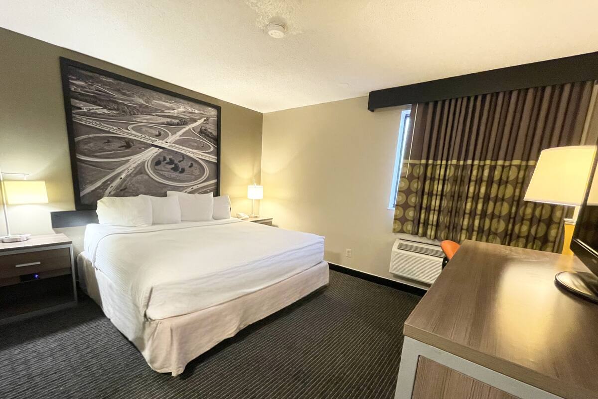 A hotel room with a large bed, bedside tables with lamps, a desk, a large abstract painting on the wall, and a window with curtains.