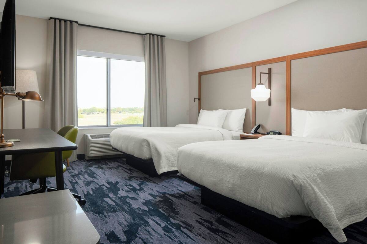 A hotel room with two beds, a desk, a chair, a lamp, and a large window with curtains.