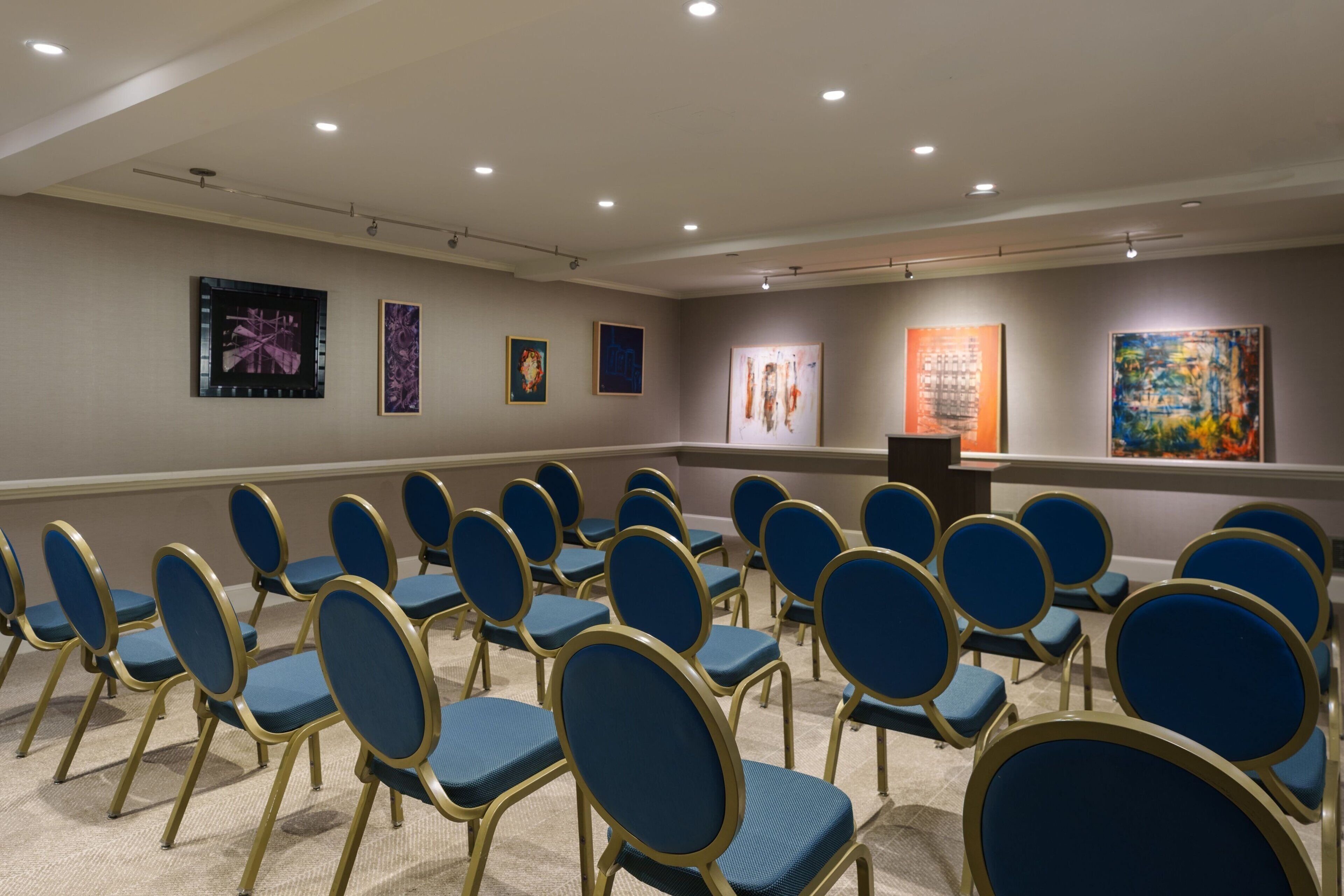 A conference room with rows of blue chairs, a podium, and several framed artworks on the walls.