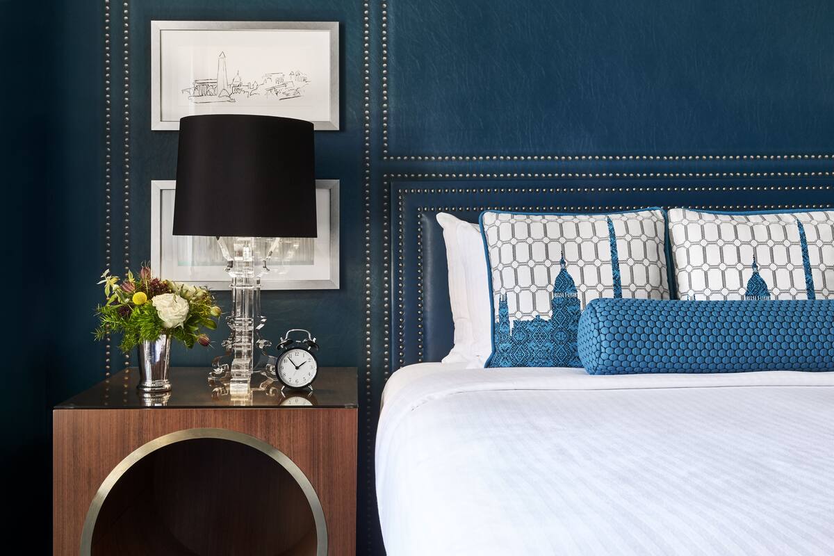 A neatly made bed with a white comforter and blue patterned pillows, a bedside table with a lamp, a framed picture, and a vase with flowers.