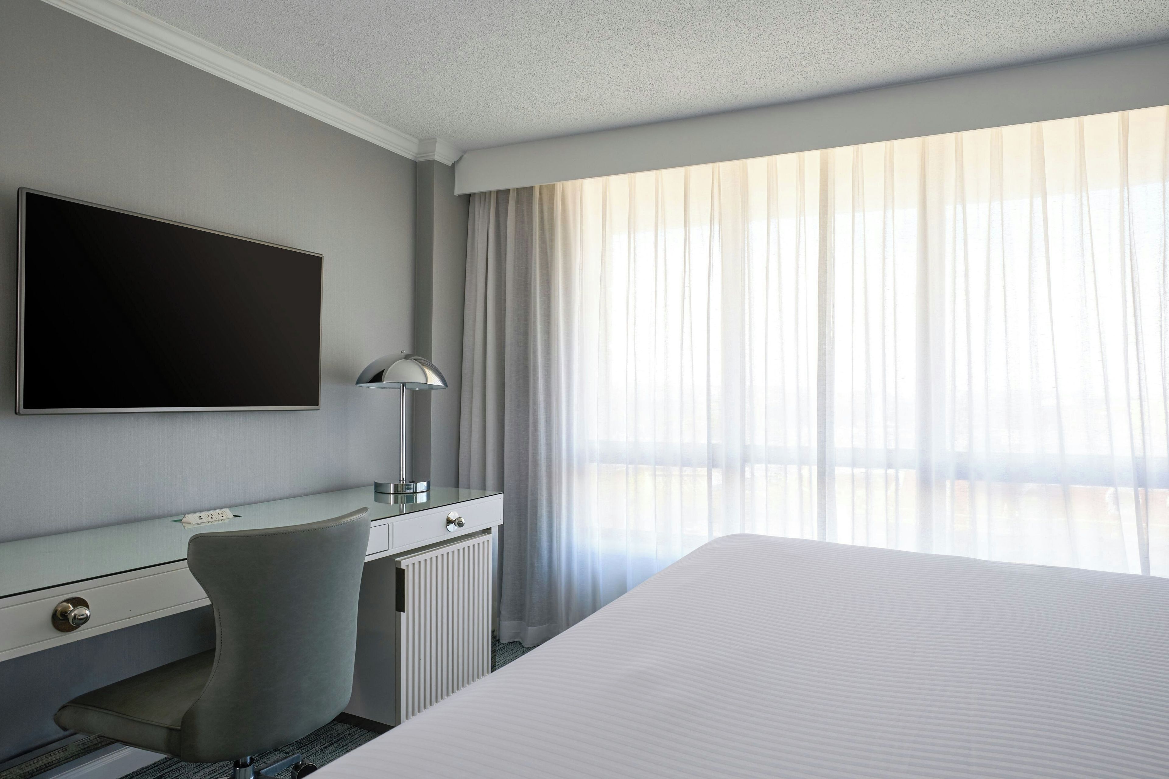 Habitació d'hotel amb llit, escriptori amb cadira, televisió i finestra amb cortines.
