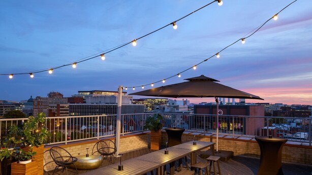 A rooftop terrace with a wooden deck, bar stools, a long table, string lights, and a large umbrella. - The Ven at Embassy Row, Washington, D.C., a Tribute Portfolio Hotel (Washington)
