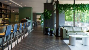 A modern bar area with a long counter and blue bar stools, a seating area with a sofa and coffee table, and a large potted plant. - The Ven at Embassy Row, Washington, D.C., a Tribute Portfolio Hotel (Washington)