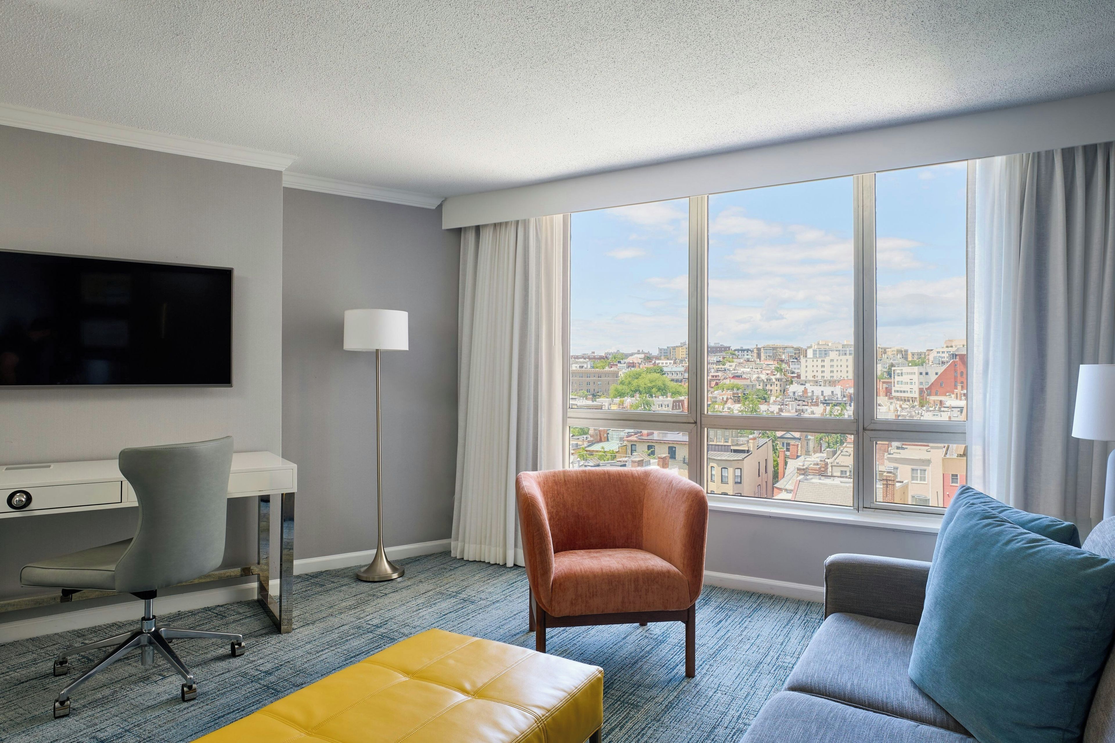 A hotel room with a desk, chair, sofa, ottoman, and a large window with a city view.