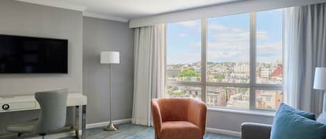 A hotel room with a desk, chair, sofa, ottoman, and a large window with a city view.