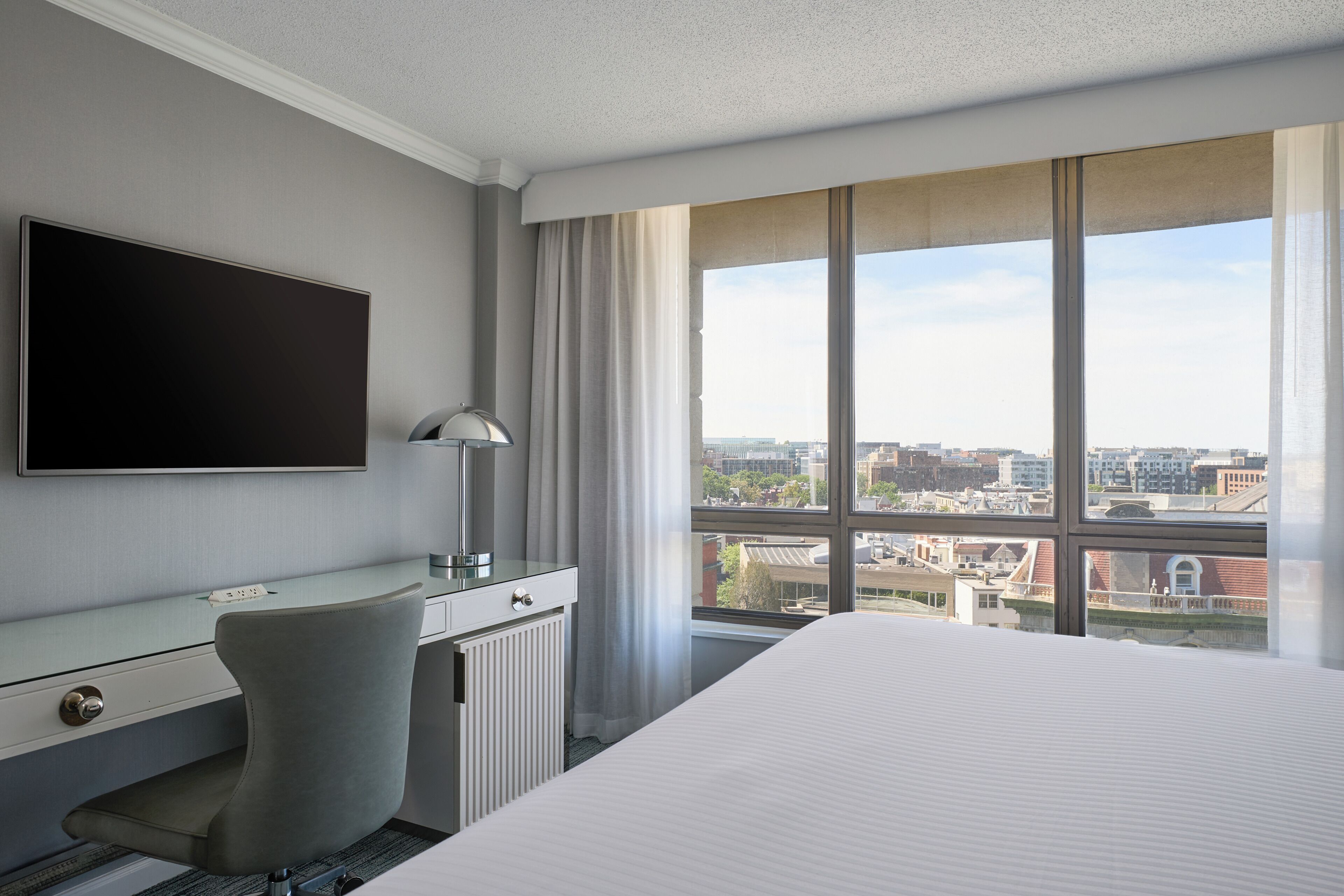 Een hotelkamer met een bed, een bureau, een stoel, een televisie en een groot raam met uitzicht op de stad.