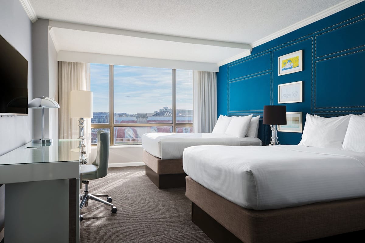 A hotel room with two beds, a desk, a chair, and a large window offering a city view.