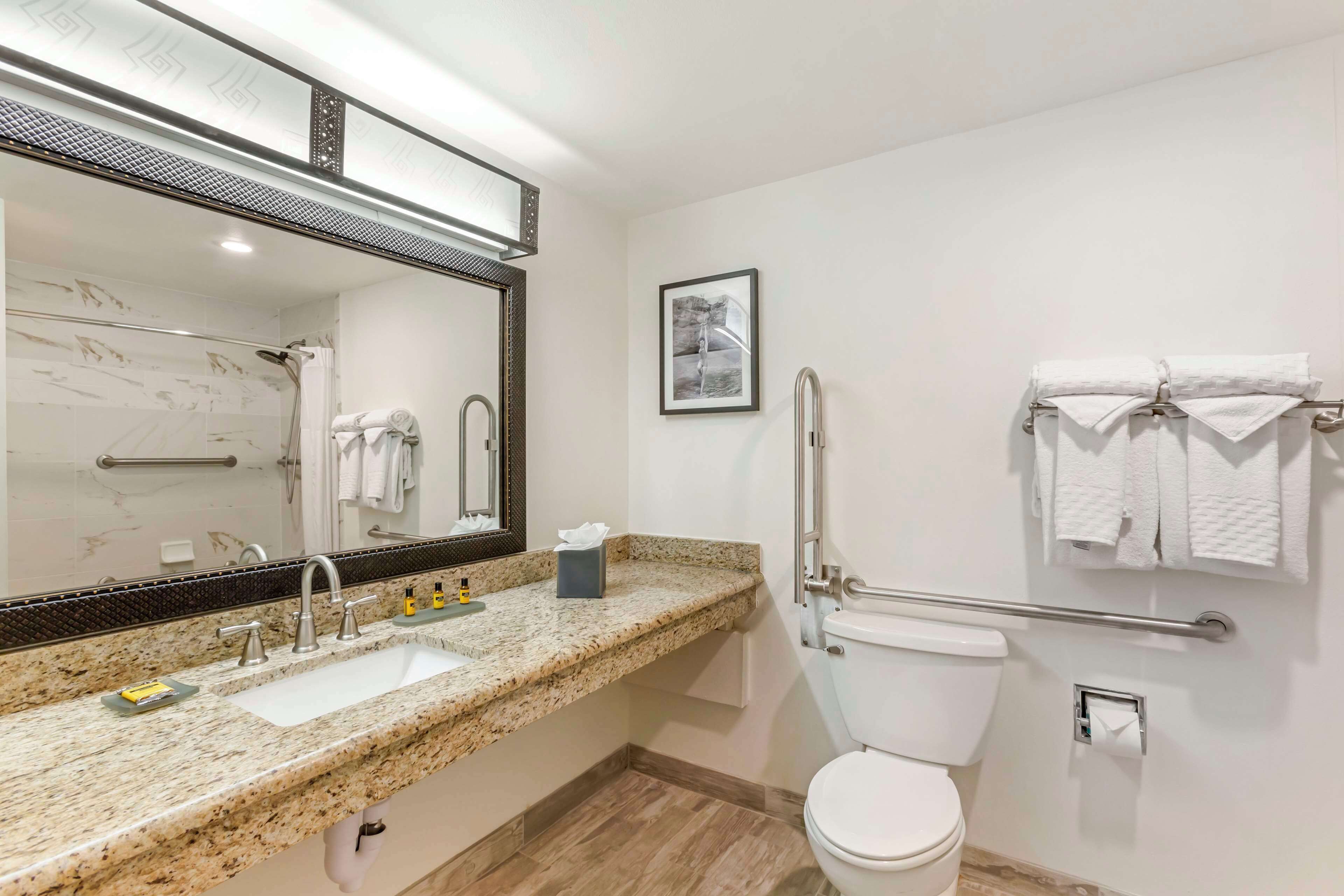 Un baño moderno con encimera de granito, un lavabo blanco, un espejo grande, un inodoro y un perchero para toallas.