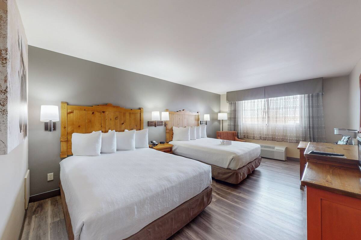 A hotel room with two beds, wooden headboards, bedside lamps, a desk, and a window with curtains.