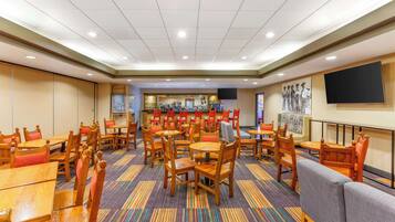 A hotel lobby with multiple wooden tables and chairs, a bar area, and a large flat-screen TV on the wall.
