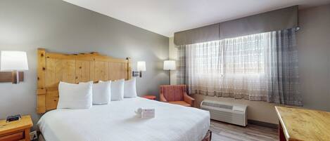Ein Hotelzimmer mit einem großen Bett, einem Holzkopfstück, einem Nachttisch, einem Sessel, einem Tisch und einem Fenster mit Vorhängen.