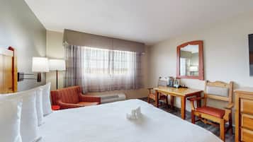 Een hotelkamer met een groot bed, een bureau met stoel, een klein tafeltje, een rode stoel, een spiegel en een raam met gordijnen.