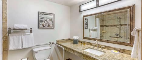 Un baño con inodoro, lavabo y espejo. Hay un perchero con toallas, un cuadro en la pared y un cubo de basura.