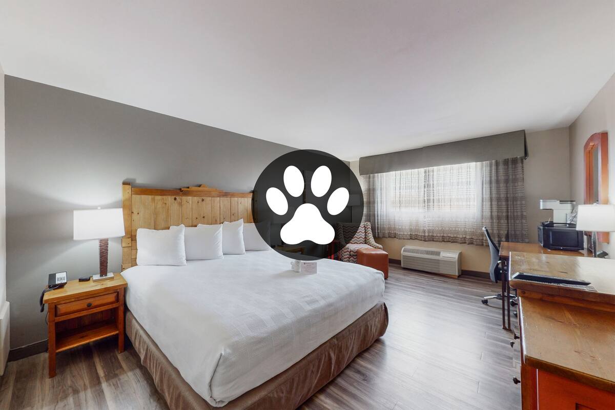 A hotel room with a large bed, a desk, a chair, a nightstand, a lamp, a telephone, and a window with curtains.